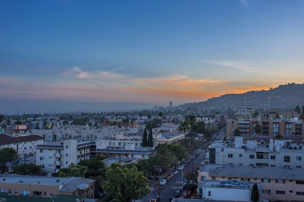 Panoramic city skyline at sunset with residential buildings, palm trees, and hillside views.