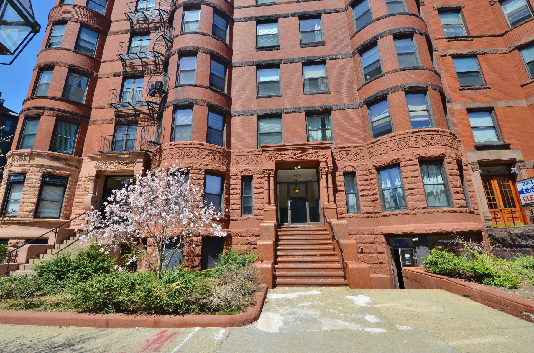 Red-brick multifamily apartment building with bay windows, landscaped entrance, and a staircase leading to the main lobby.
