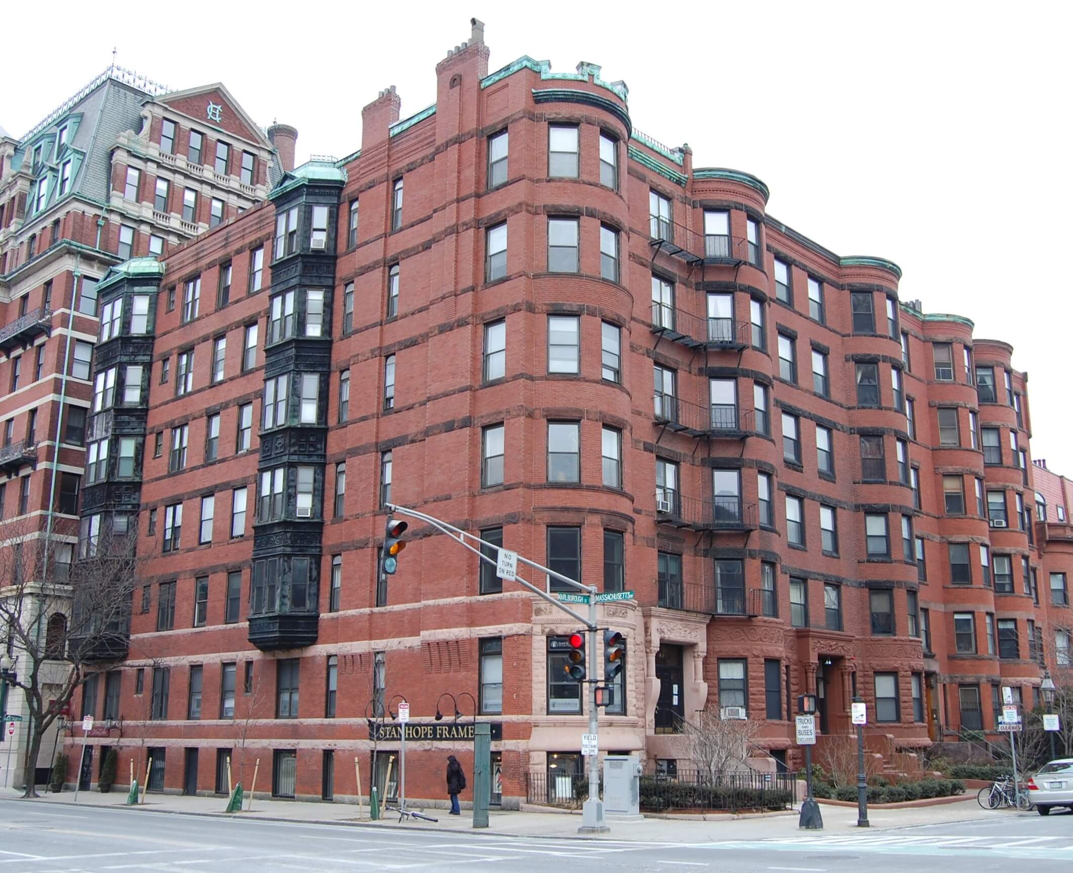 Corner view of a historic red brick apartment building at a city intersection, featuring bay windows, multiple stories, and neighboring urban buildings in the background.