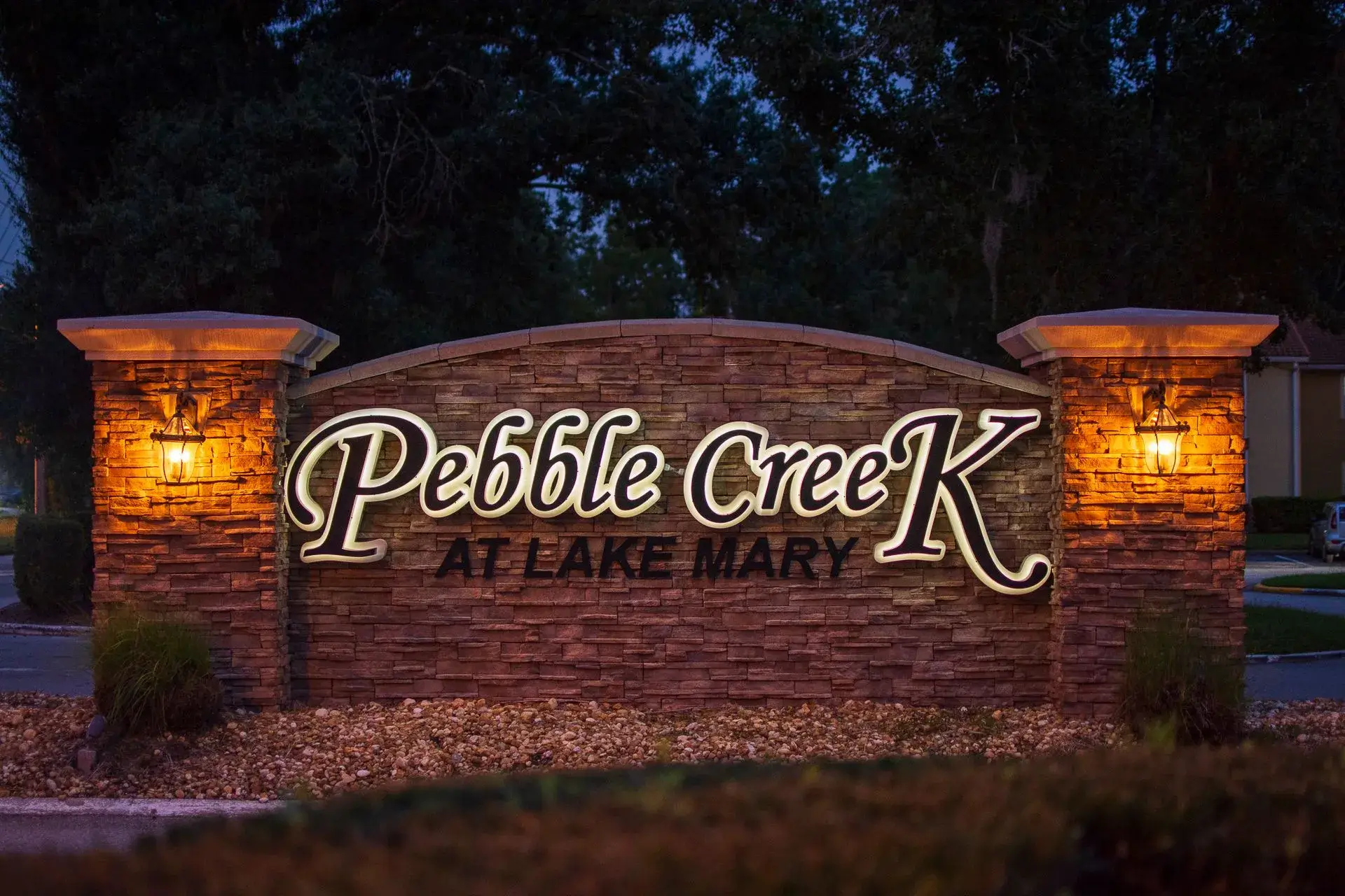 Illuminated residential community entrance sign surrounded by landscaped stone pillars and mature trees at dusk.