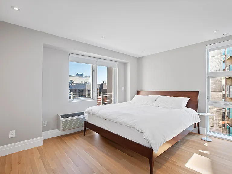 Sunlit bedroom with hardwood flooring, large windows, and minimalist furnishings.