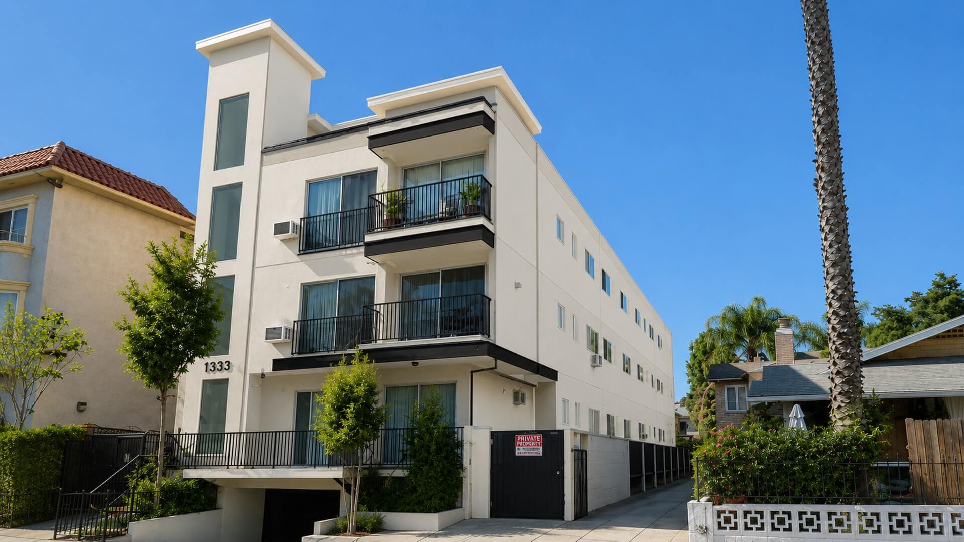 Contemporary multifamily residential building viewed from an angle, featuring private balconies, clean architectural lines, and a landscaped streetscape.