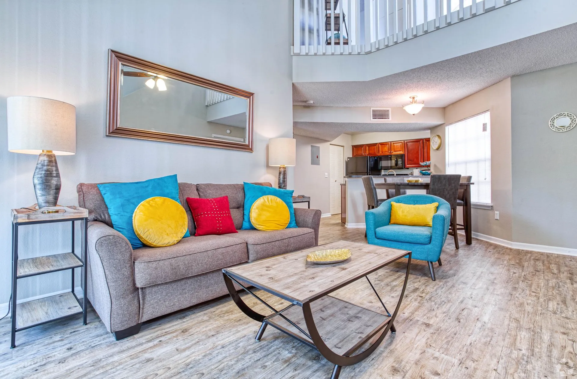Bright, modern apartment living room with a gray sofa, colorful accent pillows, glass coffee table, and open-concept dining area.