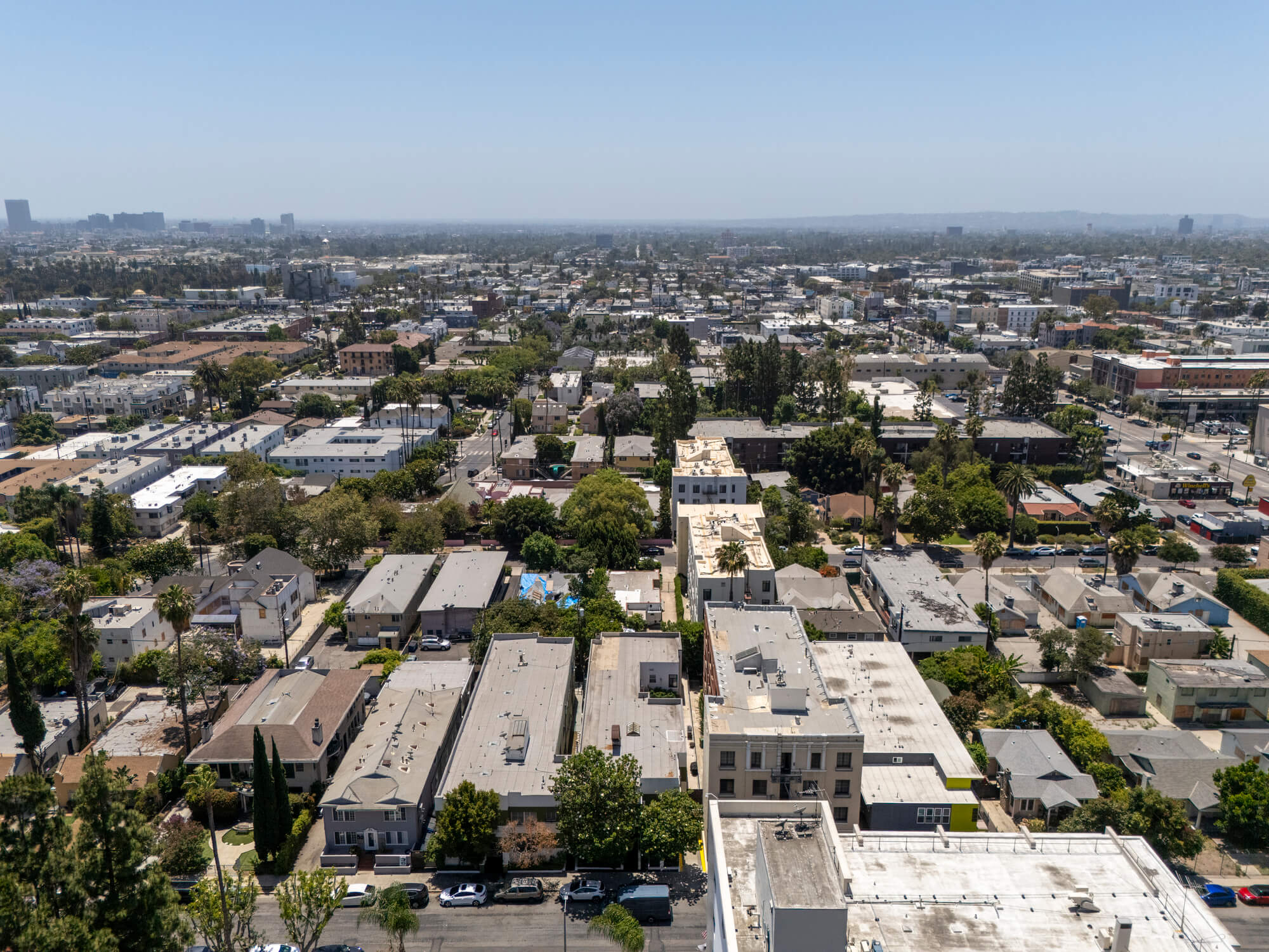Drone view of a multifamily residential neighborhood featuring apartment buildings, tree-lined streets, and surrounding urban development.