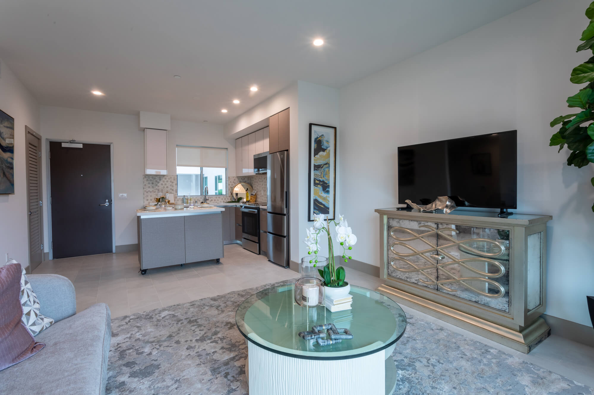 Modern apartment living room with contemporary furnishings, a wall-mounted television, and an open-concept layout connecting to the kitchen.