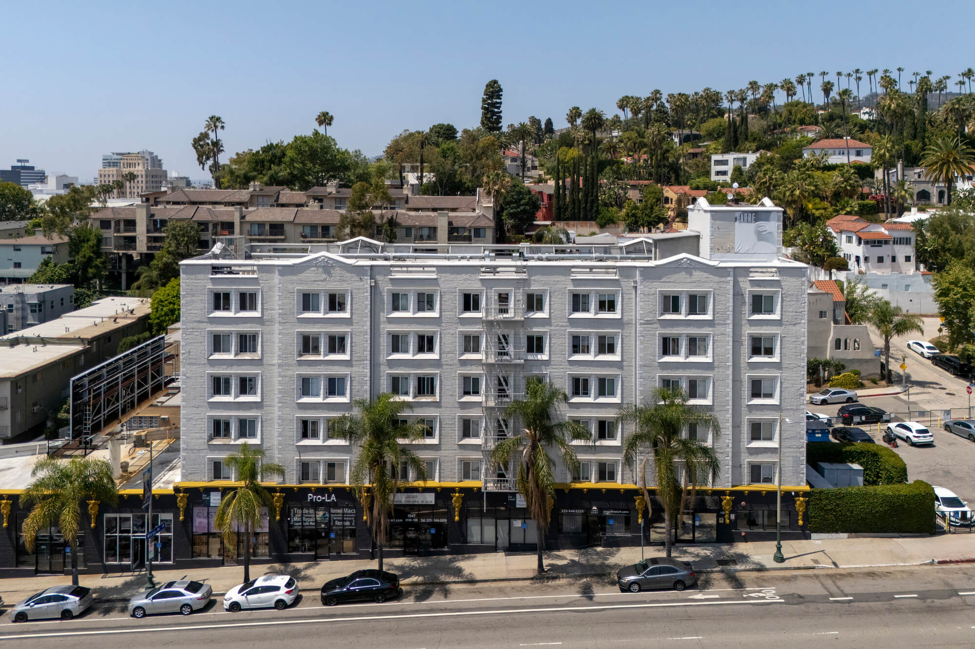 Contemporary mid-rise apartment building with a light-colored façade, street-level retail space, and landscaped frontage along a major boulevard.