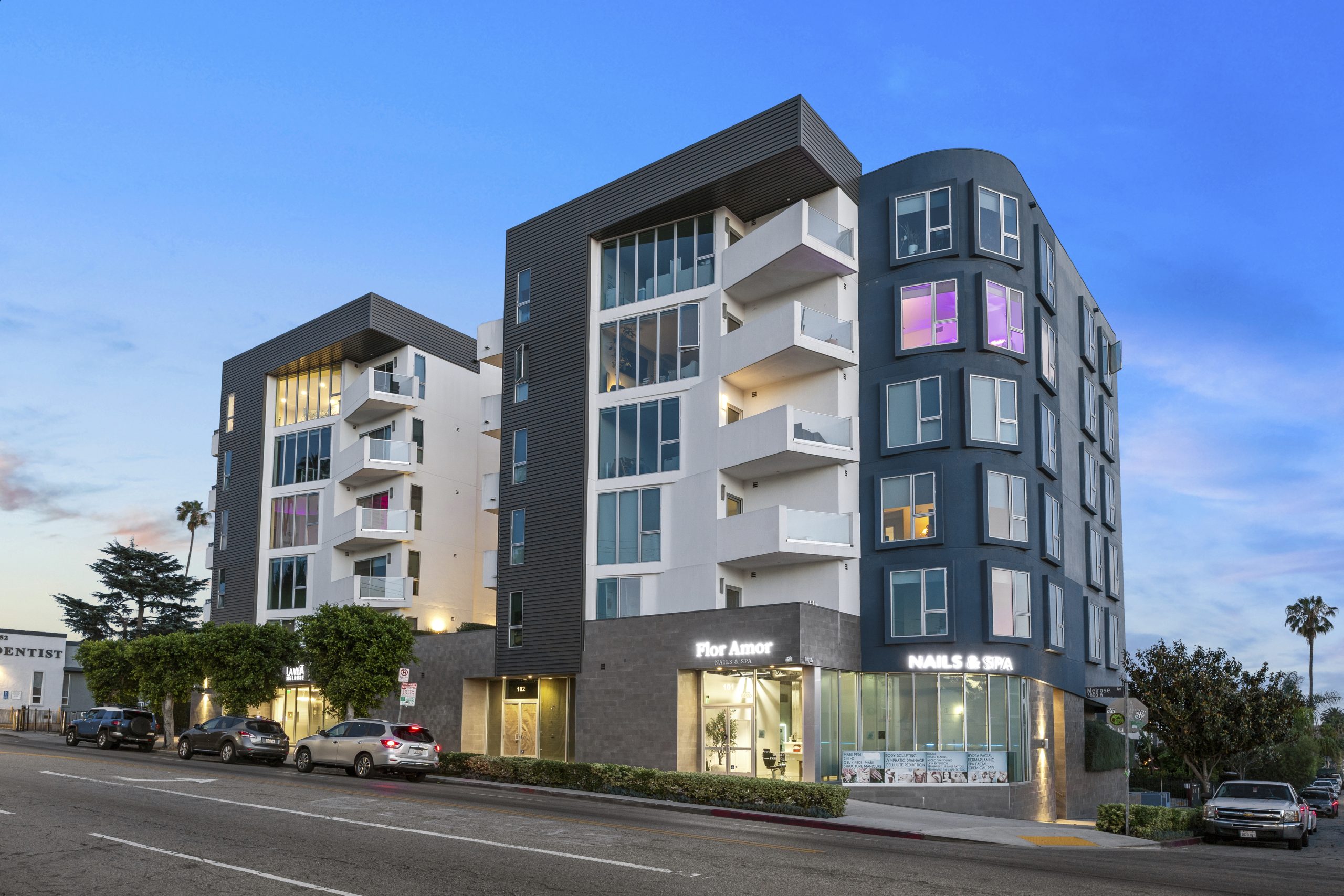 Modern six-story apartment building with contemporary architecture, featuring private balconies, large windows, ground-floor retail space, and evening exterior lighting.