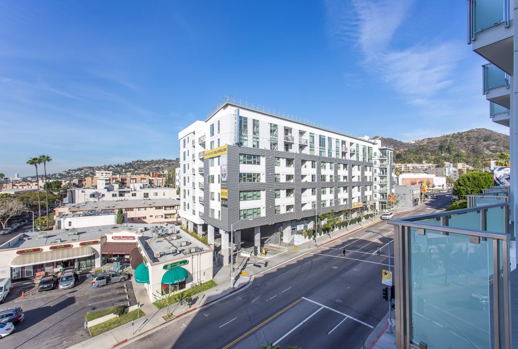 Modern multifamily apartment building with balconies overlooking urban street and hillside skyline.