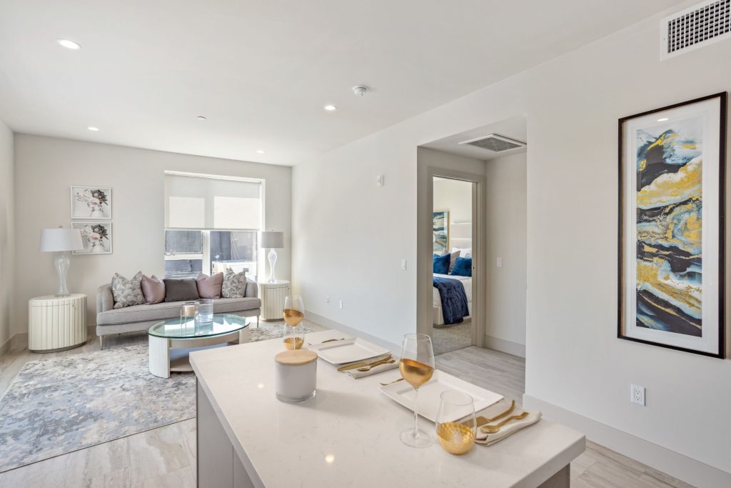 A bright open-concept apartment featuring a modern kitchen island with place settings, a cozy living area with a sofa and coffee table, and a bedroom visible through an open doorway.