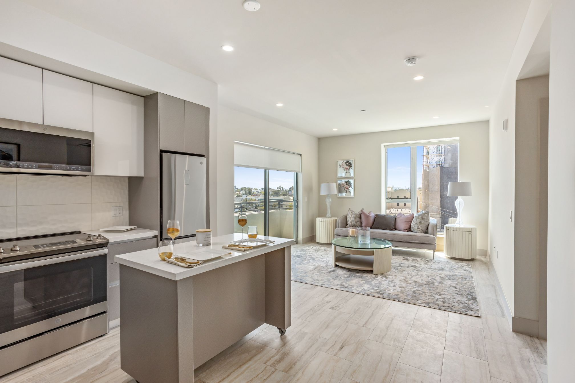 A modern open-concept living space featuring a white kitchen with a center island, stainless steel appliances, and a bright living room with large windows, neutral furnishings, and a cozy seating area.