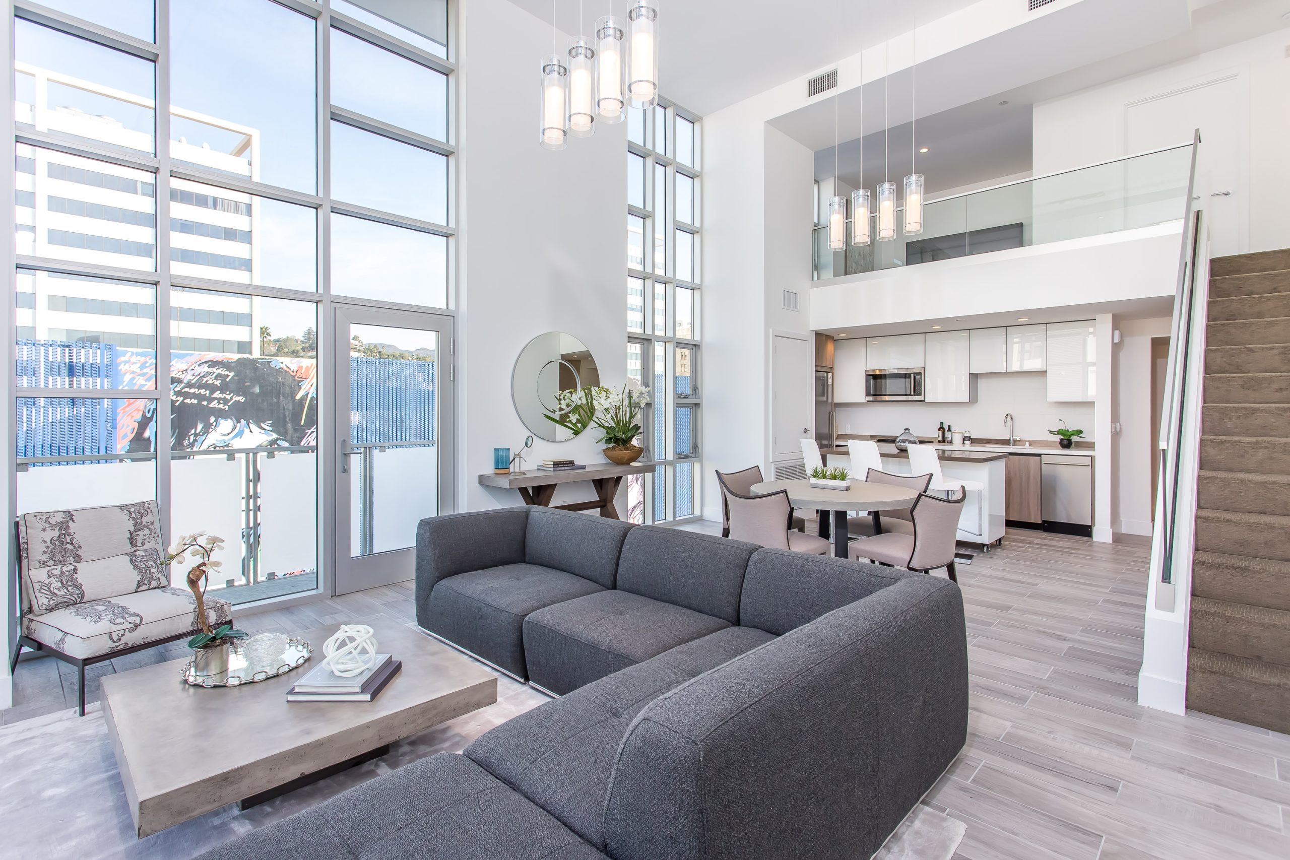 A bright open-concept living room and kitchen with high ceilings, large windows, modern furnishings, and abundant natural light.