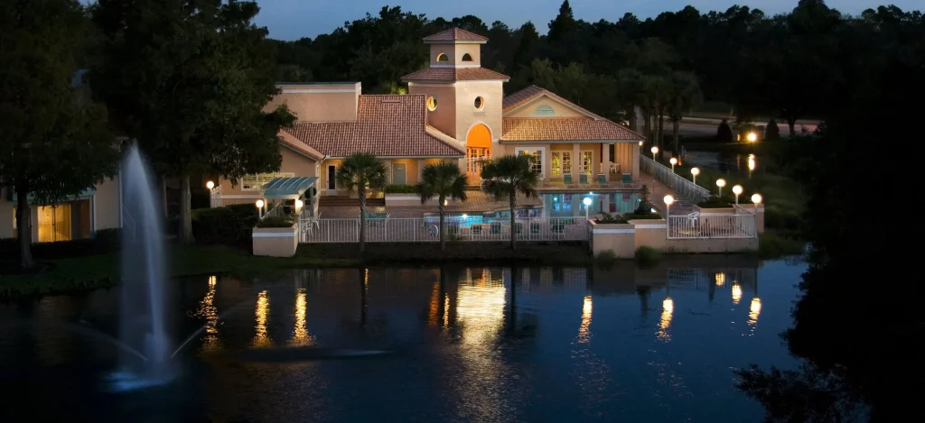 A lakeside community clubhouse at dusk featuring a lit swimming pool, palm trees, and a fountain reflecting on the water.